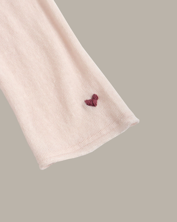 A close-up of a light pink ribbed knit fabric sleeve or cuff with a small embroidered burgundy heart near the cuff edge, set against a solid taupe background.