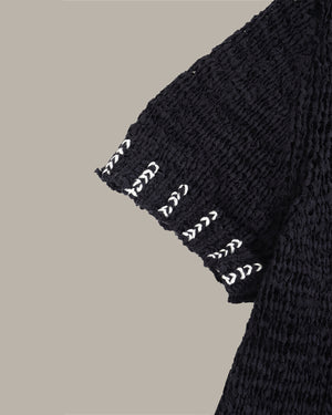 Close-up of a black textured knit garment sleeve with white embroidered chevron patterns along the hem, set against a solid beige background.