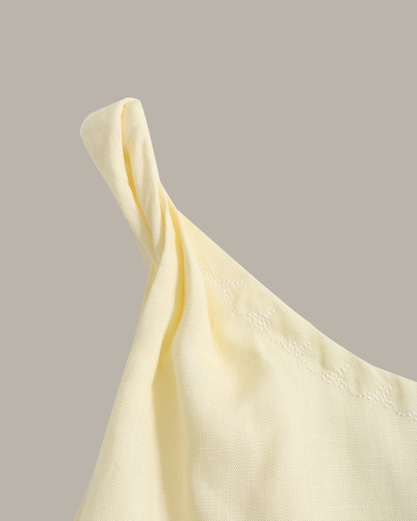 A close-up of a pale yellow fabric garment with a gathered shoulder and subtle embroidered detailing along the neckline, set against a solid taupe background.