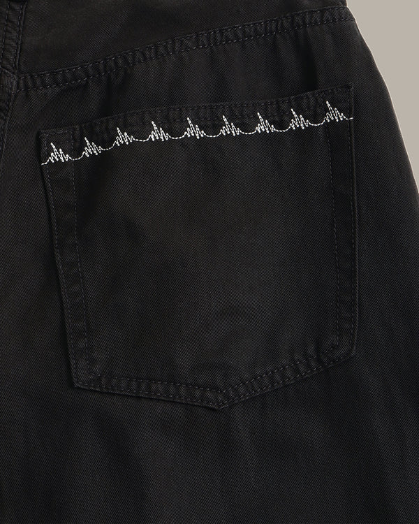 Close-up of a black denim pocket with a white embroidered heartbeat line across the top, set against a neutral gray background.