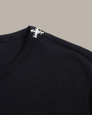 Close-up of a black crewneck t-shirt featuring a white embroidered cross logo on the inside of the collar, set against a solid taupe background.