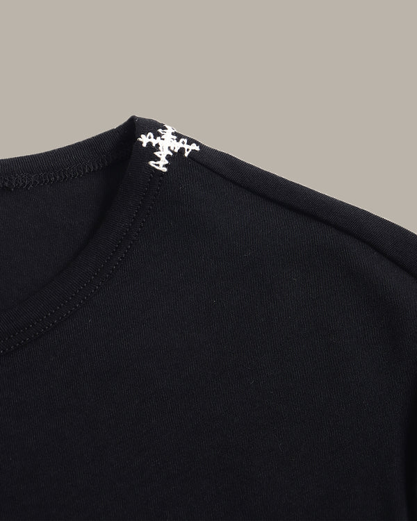 Close-up of a black crewneck t-shirt featuring a white embroidered cross logo on the inside of the collar, set against a solid taupe background.
