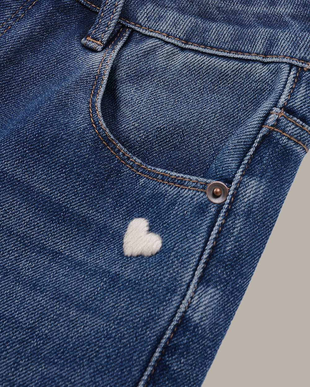 A pair of medium-wash blue denim jeans with a slight flare, featuring a small white embroidered heart on the right pocket and a small white embroidered logo on the left thigh, shown against a solid taupe background.