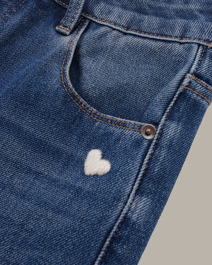 Close-up of a pair of blue denim jeans showing the front pocket with a small white embroidered heart detail.