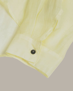 Close-up of a pale yellow button-down shirt cuff with a single dark button, set against a solid taupe-gray background.