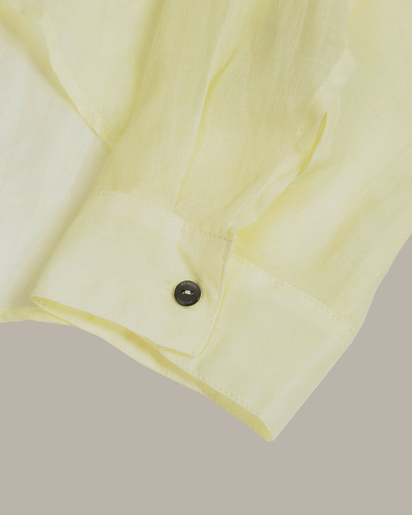 Close-up of a pale yellow button-down shirt cuff with a single dark button, set against a solid taupe-gray background.