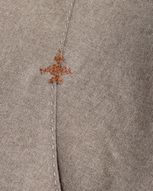 Close-up of a taupe-brown suede or nubuck fabric with a central vertical seam and a small, decorative rust-brown embroidered motif near the top.