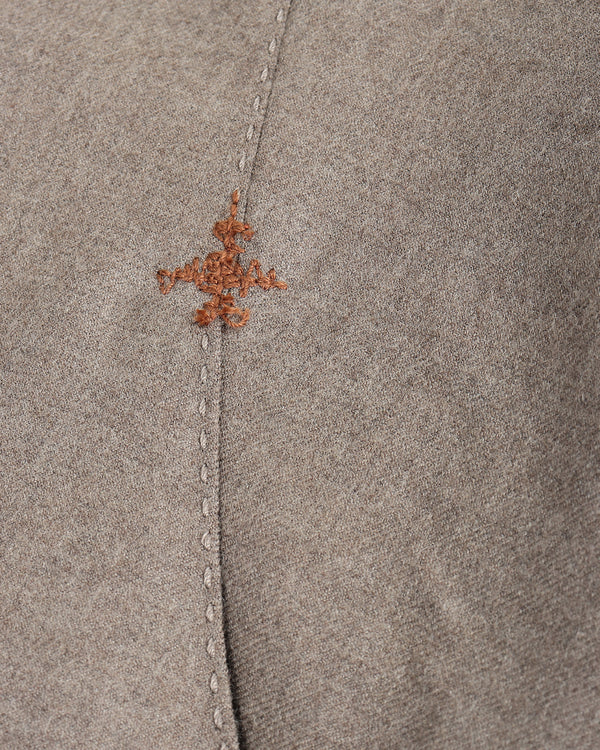Close-up of a taupe-brown suede or nubuck fabric with a central vertical seam and a small, decorative rust-brown embroidered motif near the top.