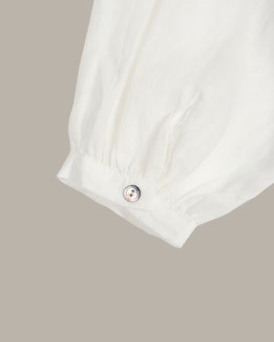Close-up of a white fabric sleeve or cuff with a gathered elastic band and a small round metallic button, shown against a plain warm gray background.