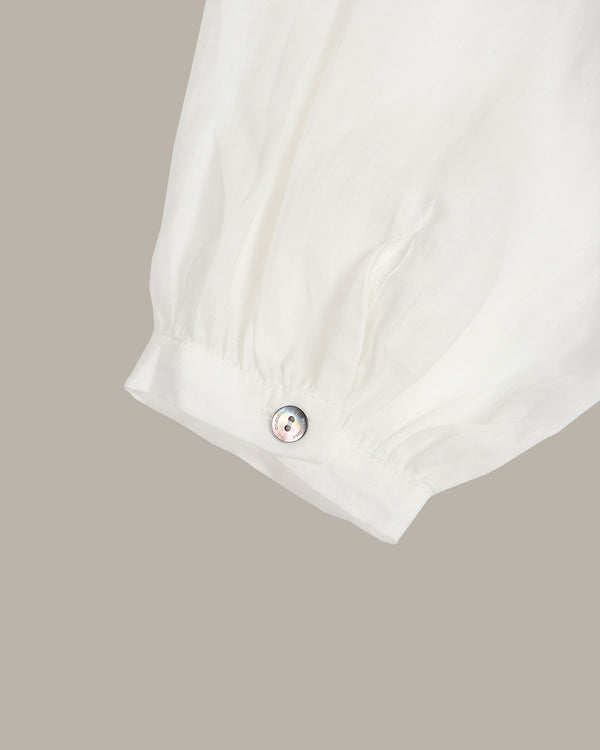Close-up of a white fabric sleeve or cuff with a gathered elastic band and a small round metallic button, shown against a plain warm gray background.