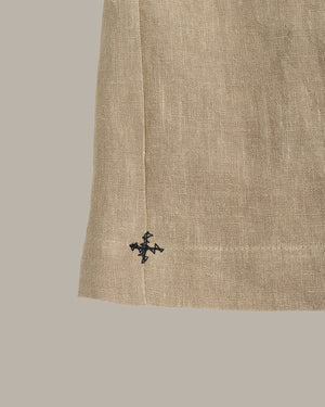 Close-up of a beige linen fabric garment corner with a small black embroidered cross logo near the hem, set against a solid taupe background.