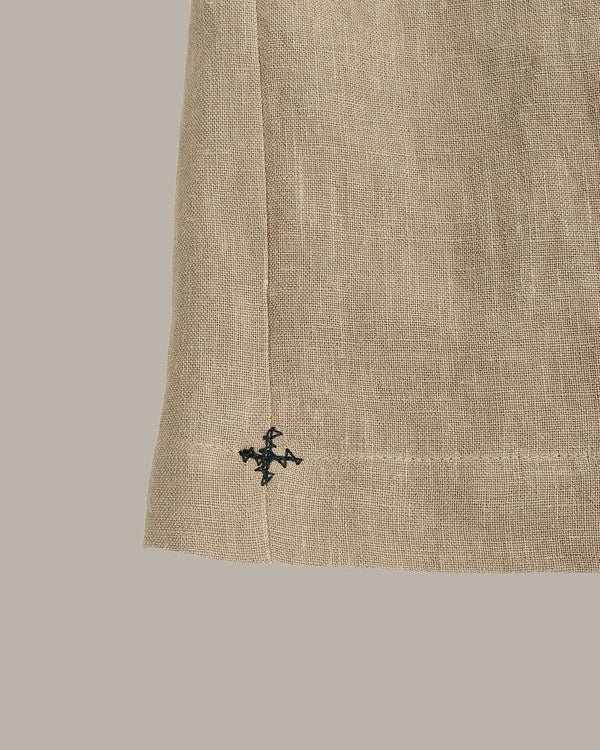 Close-up of a beige linen fabric garment corner with a small black embroidered cross logo near the hem, set against a solid taupe background.