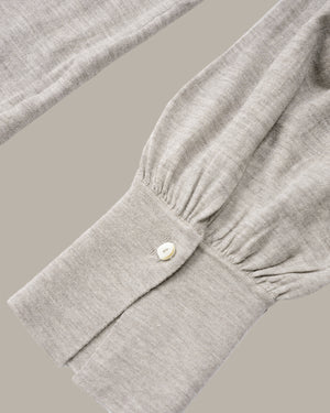 Close-up of a light gray linen-blend long-sleeve top showing the gathered cuff and a single button closure, set against a solid taupe background.
