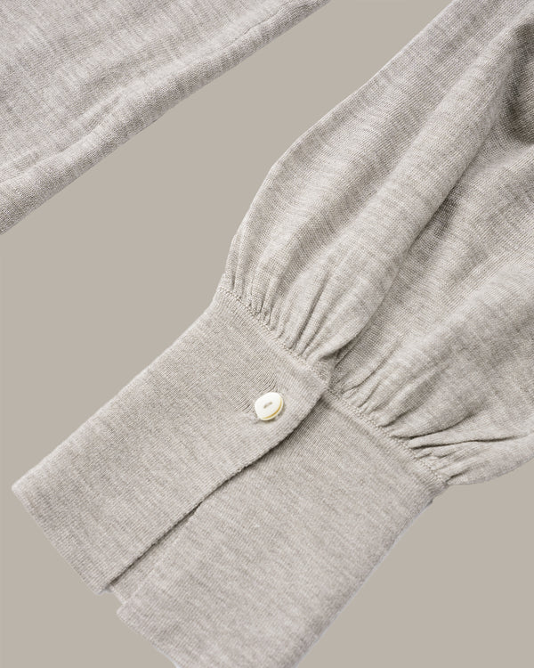 Close-up of a light gray linen-blend long-sleeve top showing the gathered cuff and a single button closure, set against a solid taupe background.
