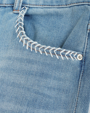 Close-up of a light blue denim pocket with decorative white zigzag stitching around the pocket opening and a silver metal rivet.