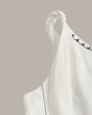 Close-up of a white sleeveless top with a twisted shoulder strap and black script embroidery along the neckline, set against a solid taupe background.