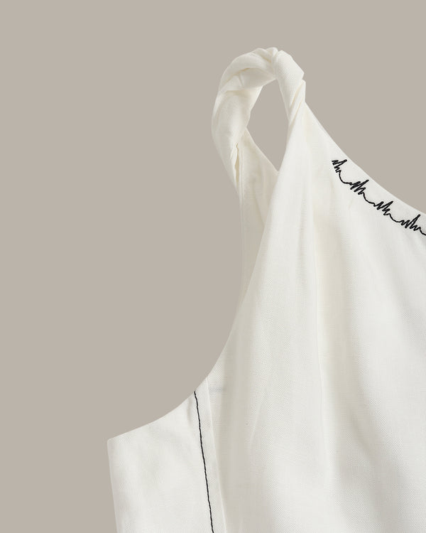 Close-up of a white sleeveless top with a twisted shoulder strap and black script embroidery along the neckline, set against a solid taupe background.