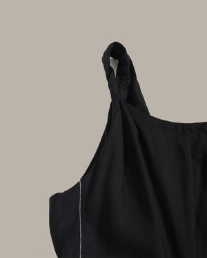 Close-up of a black fabric garment with a twisted shoulder strap detail and white contrast stitching, shown against a solid taupe background.