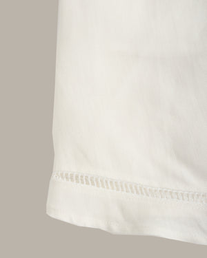 Close-up of a white fabric garment showing a folded hem with a decorative perforated or eyelet stitched edge, against a plain warm gray background.