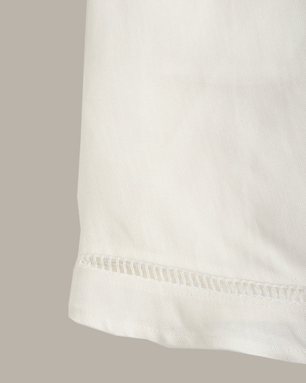 Close-up of a white fabric garment showing a folded hem with a decorative perforated or eyelet stitched edge, against a plain warm gray background.