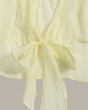 A close-up of a pale yellow or butter yellow fabric garment with a tied bow detail at the waist, showing the soft texture and drape of the material against a neutral light gray background.
