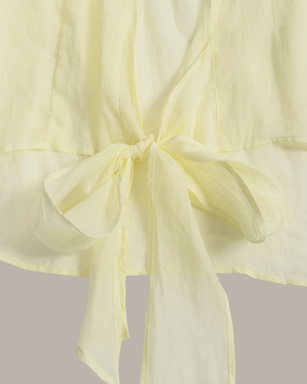 A close-up of a pale yellow or butter yellow fabric garment with a tied bow detail at the waist, showing the soft texture and drape of the material against a neutral light gray background.
