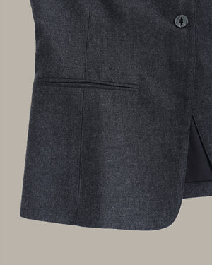 Close-up of a dark charcoal gray textured blazer fabric, showing a button, pocket seam, and side slit against a solid beige background.