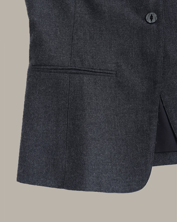 Close-up of a dark charcoal gray textured blazer fabric, showing a button, pocket seam, and side slit against a solid beige background.