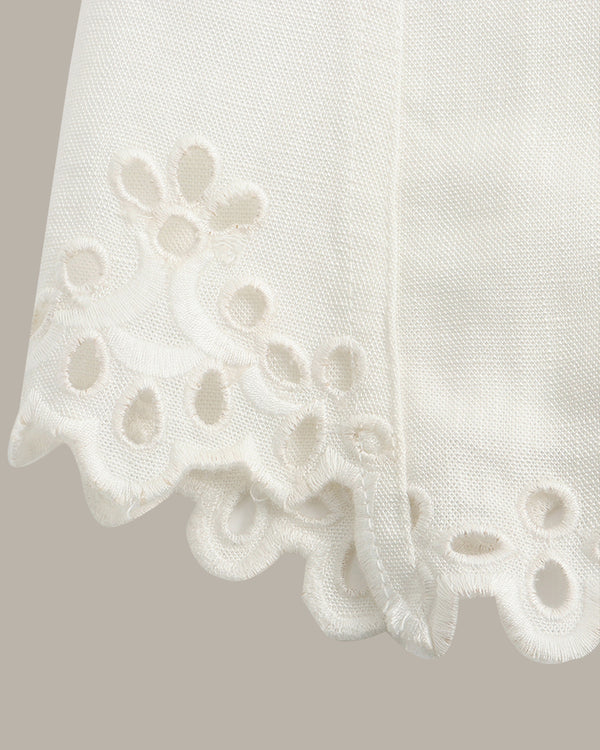 Close-up of a white eyelet fabric with a scalloped edge and floral cutwork pattern, shown against a plain warm gray background.