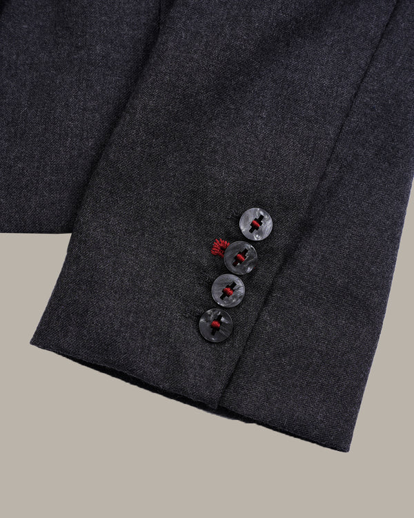 Close-up of a dark charcoal gray wool suit jacket sleeve, showing three dark buttons with red centers on the cuff, set against a solid beige background.