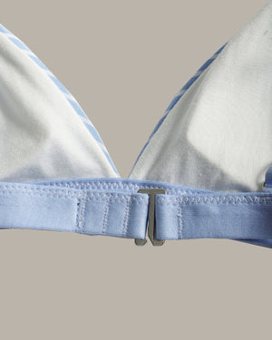 Close-up of a light blue and white triangle bralette or bikini top, showing the underband, silver metal clasp, and striped fabric edge against a solid beige background.