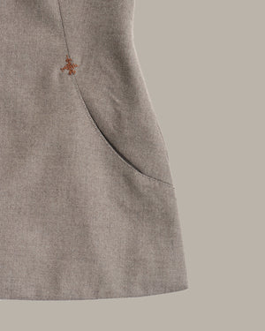 Close-up of a light taupe-gray textured fabric garment with a small orange embroidered logo and a curved seam detail, set against a solid taupe background.