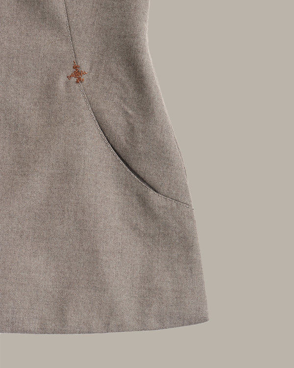 Close-up of a light taupe-gray textured fabric garment with a small orange embroidered logo and a curved seam detail, set against a solid taupe background.