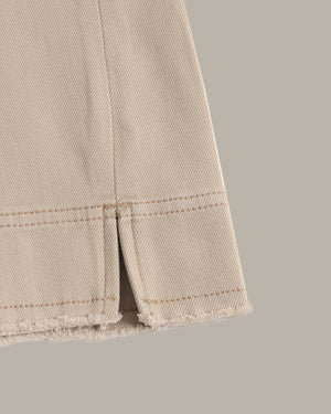 Close-up of a beige denim fabric detail showing a frayed hem and a vertical slit with neat stitching, set against a solid taupe background.