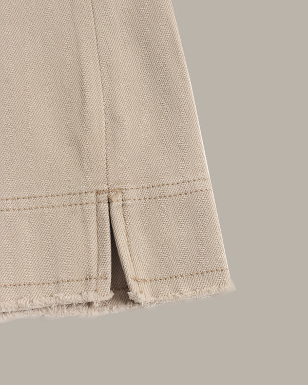 Close-up of a beige denim fabric detail showing a frayed hem and a vertical slit with neat stitching, set against a solid taupe background.