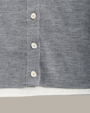 Close-up of a gray ribbed knit cardigan or sweater with a vertical button placket and white buttons, showing a white ribbed inner hem layer and a neutral beige background.