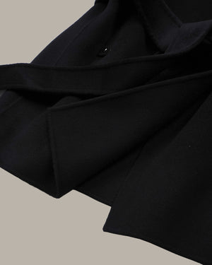 Close-up detail shot of a black coat or jacket with a wide lapel and a small dark button, set against a solid warm gray background.