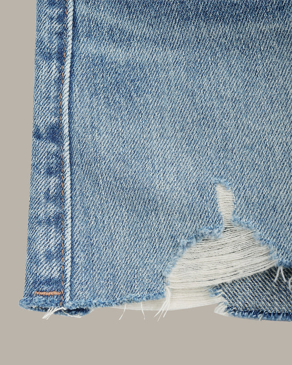 Close-up of light blue denim shorts showing a distressed raw hem with frayed white thread and orange contrast stitching, set against a solid taupe background.