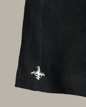 Close-up of a black textured fabric item, likely a skirt or drape, featuring a small white embroidered logo near the lower hem against a solid taupe background.