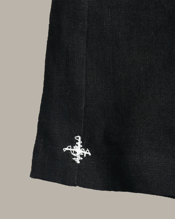 Close-up of a black textured fabric item, likely a skirt or drape, featuring a small white embroidered logo near the lower hem against a solid taupe background.