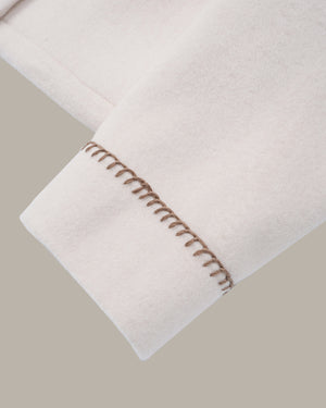 Close-up of a light cream or off-white fabric cuff with decorative brown contrast stitching, shown against a solid taupe-gray background.