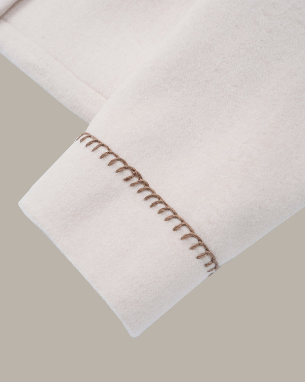 Close-up of a light cream or off-white fabric cuff with decorative brown contrast stitching, shown against a solid taupe-gray background.