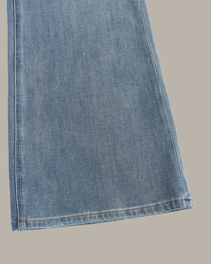 A close-up of a light blue denim fabric leg, showing the texture and hem stitching, set against a solid taupe background.