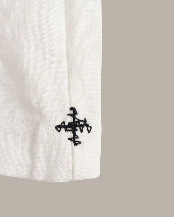 Close-up of a white textured fabric sleeve with a black embroidered abstract cross-like logo near the cuff, set against a solid taupe background.