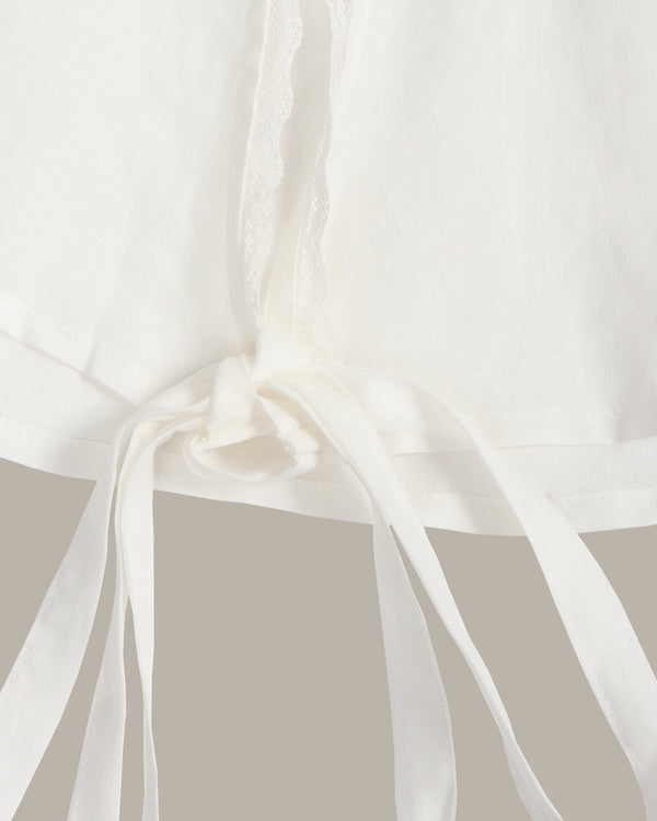 Close-up of a white fabric garment with a tie detail and soft draped ribbon, shown against a plain light gray background.