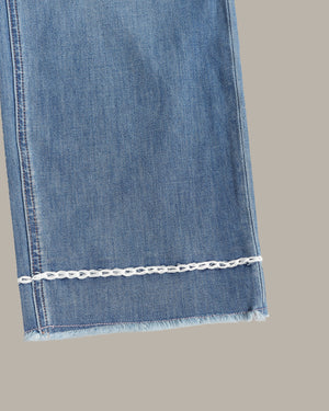 Close-up of a blue denim fabric leg with a frayed hem and a decorative white chain detail running horizontally near the cuff, shown against a plain taupe background.