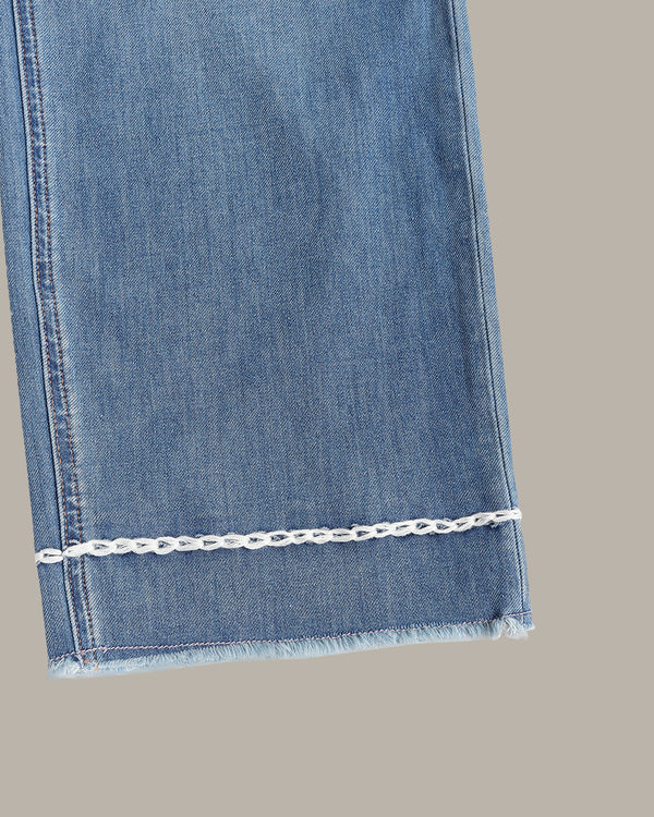 Close-up of a blue denim fabric leg with a frayed hem and a decorative white chain detail running horizontally near the cuff, shown against a plain taupe background.